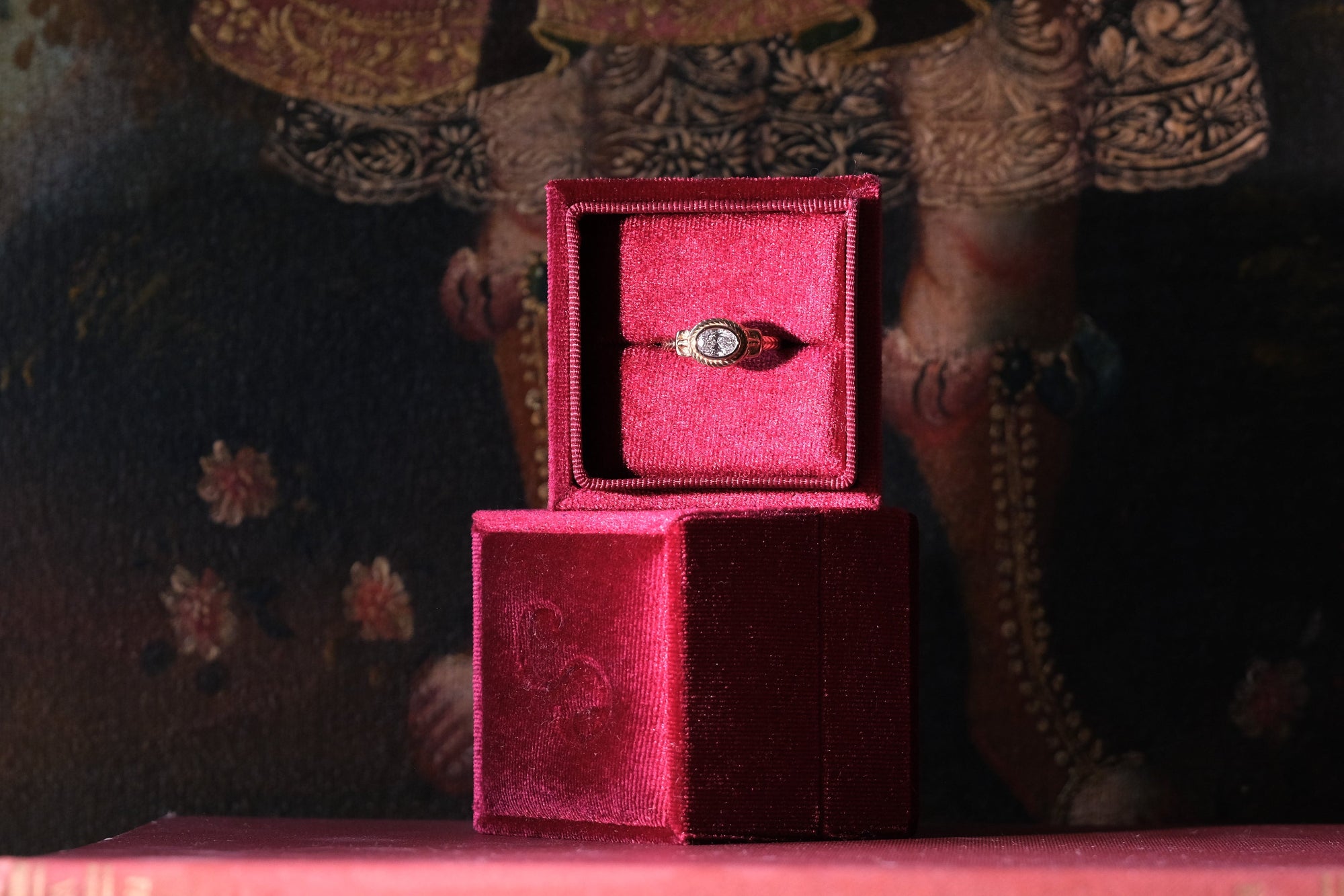 Red velvet jewelry box with a ring on top of a book against an ornate background